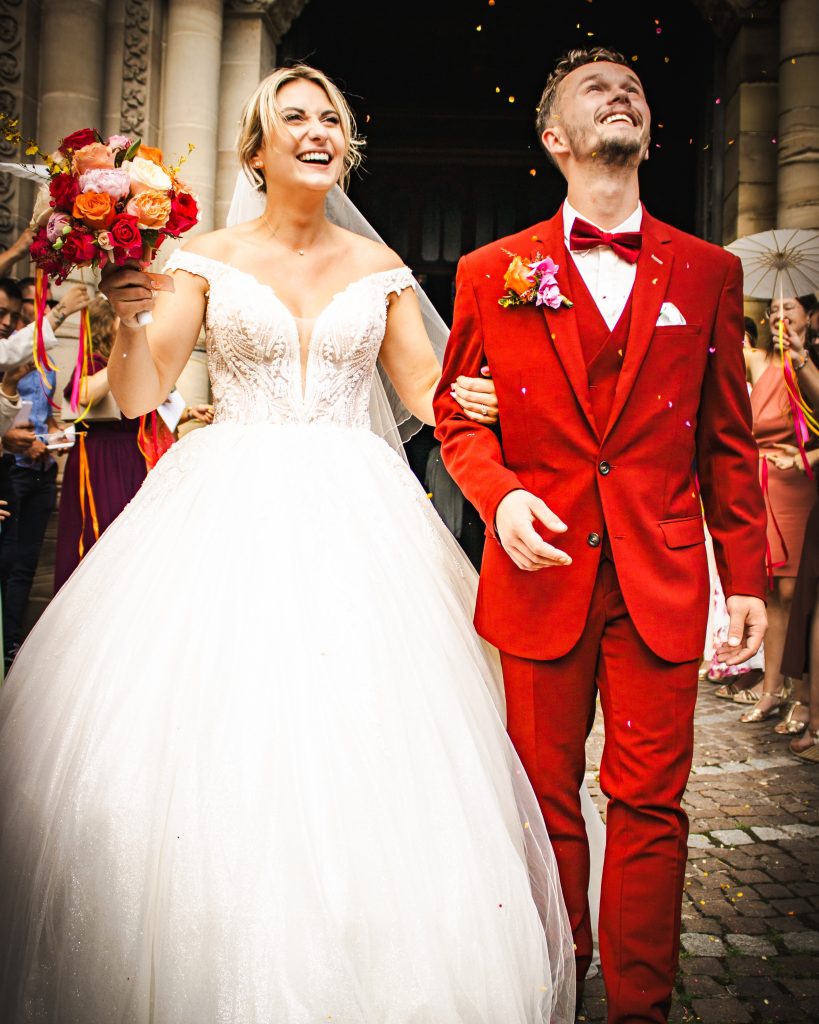 Couple de mariés marchant dans l’allée, la mariée en robe blanche et le marié en costume rouge