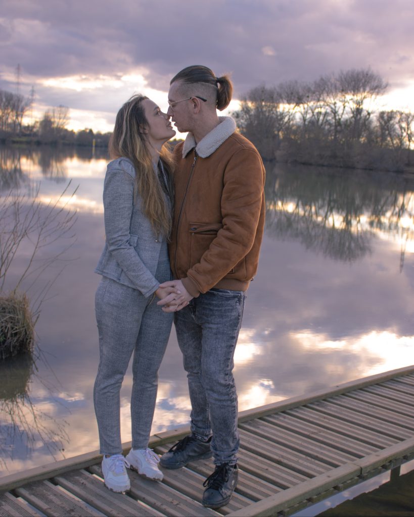 Couple s’enlaçant sur un pont en bois au bord de l’eau, au coucher du soleil.